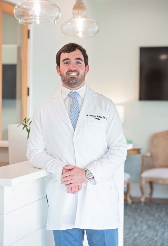 Dr. Sandy Vallee of Point Clear Dental Associates in Fairhope, AL. They are leaning against a white counter with hands clasped, while the background features a chair, a table with a plant, and a mounted television in a professional office setting.