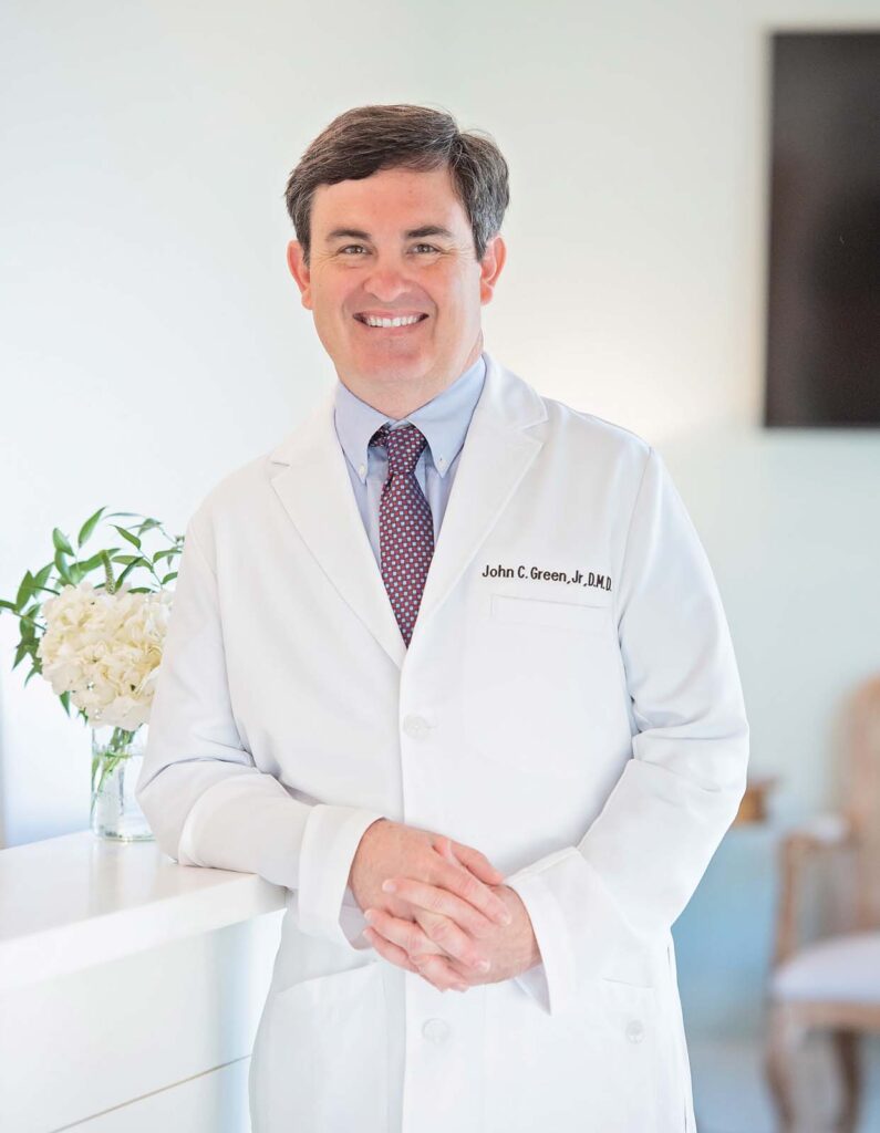 Dr. John Green of Point Clear Dental Associates. He stand with hands clasped in front of a white counter, which holds a vase with white flowers, while a chair and a television screen are visible in the blurred background.