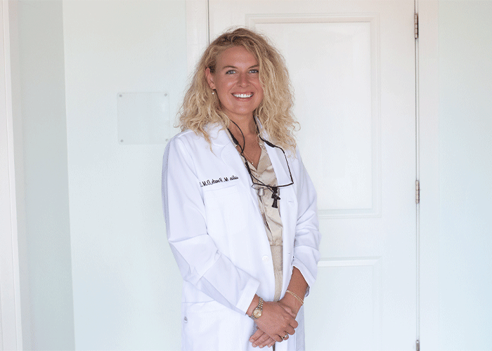 Headshot of Dr. Kalin Roach of Point Clear Dental Associates in Fairhope, AL. She is wearing a white lab coat, standing in front of a white door. They have curly blonde hair, wear a beige blouse underneath, and accessorize with a cross pendant necklace, glasses hanging around their neck, a watch, and a bracelet, reflecting a professional, likely medical, setting.