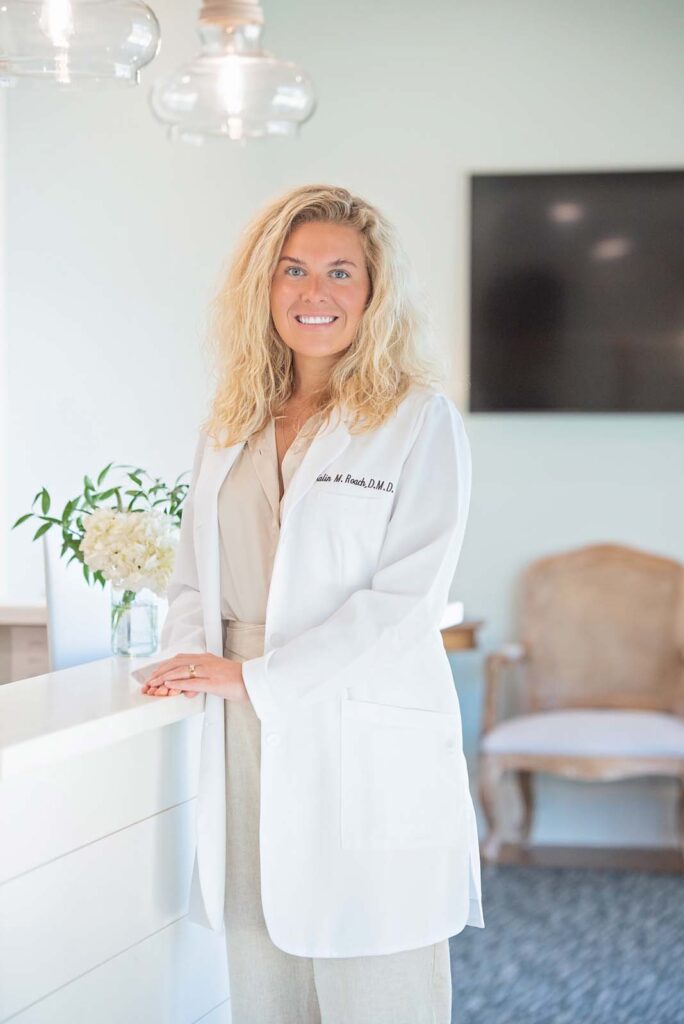 Dr. Kalin McCabe Roach of Point Clear Dental Associates. The person is standing next to a white counter with a vase of white flowers on it. In the background, there is a chair and a large flat-screen television mounted on the wall.