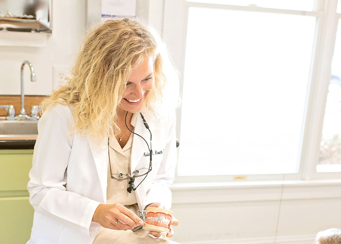 Dr. Roach of Point Clear Dental Associates in Fairhope, AL in a white lab coat holding a dental model and a dental tool, likely demonstrating or explaining dental care. The bright background includes a sink and a window, emphasizing a professional setting.