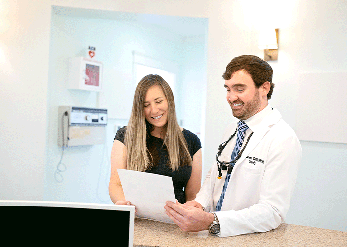 Dr. Sandy Vallee of Point Clear Dental Associates in Fairhope, AL standing next to a woman. The medical professional is holding a piece of paper and appears to be discussing its contents with the woman. The setting is a medical office or clinic, indicated by medical equipment on the wall, including an AED (Automated External Defibrillator) and other devices.