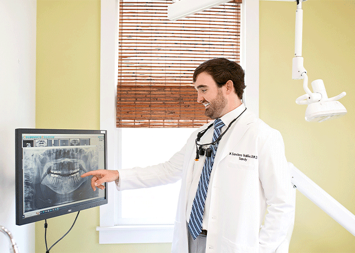 Dr. Sandy Vallee of Point Clear Dental Associates in Fairhope, AL wearing a white lab coat, pointing at a computer screen displaying multiple dental X-rays. The setting includes a window with a bamboo shade, a dental light fixture, and various dental equipment, highlighting a clinical environment focused on diagnostics.