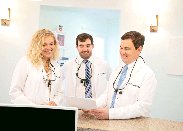 Drs. Roach, Vallee, and Green of Point Clear Dental Associates in Fairhope, AL. The image features three healthcare professionals in white lab coats gathered around a counter in a medical or dental office. One holds a clipboard, while an AED sign is visible in the background, highlighting a professional and collaborative healthcare setting.