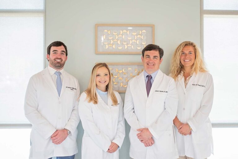 The doctors at Point Clear Dental Associates, LLC in Fairhope, AL. The image features four individuals standing side by side, all wearing white lab coats with names embroidered on them. They are positioned in front of a wall adorned with two framed artworks displaying golden leaf patterns, suggesting a professional setting.