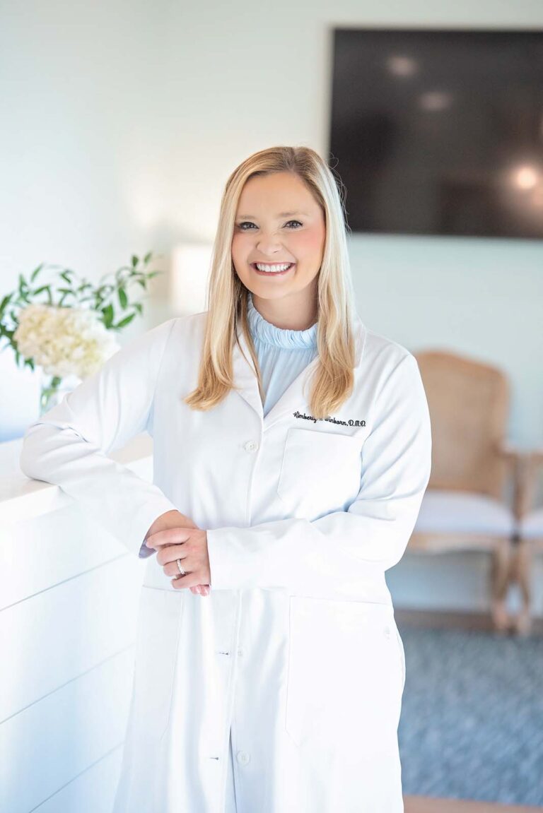 Dr. Winborn of Point Clear Dental Associates, standing in a professional setting. They lean against a white surface with a vase of white flowers beside them, while a chair and a mounted television screen are visible in the background.