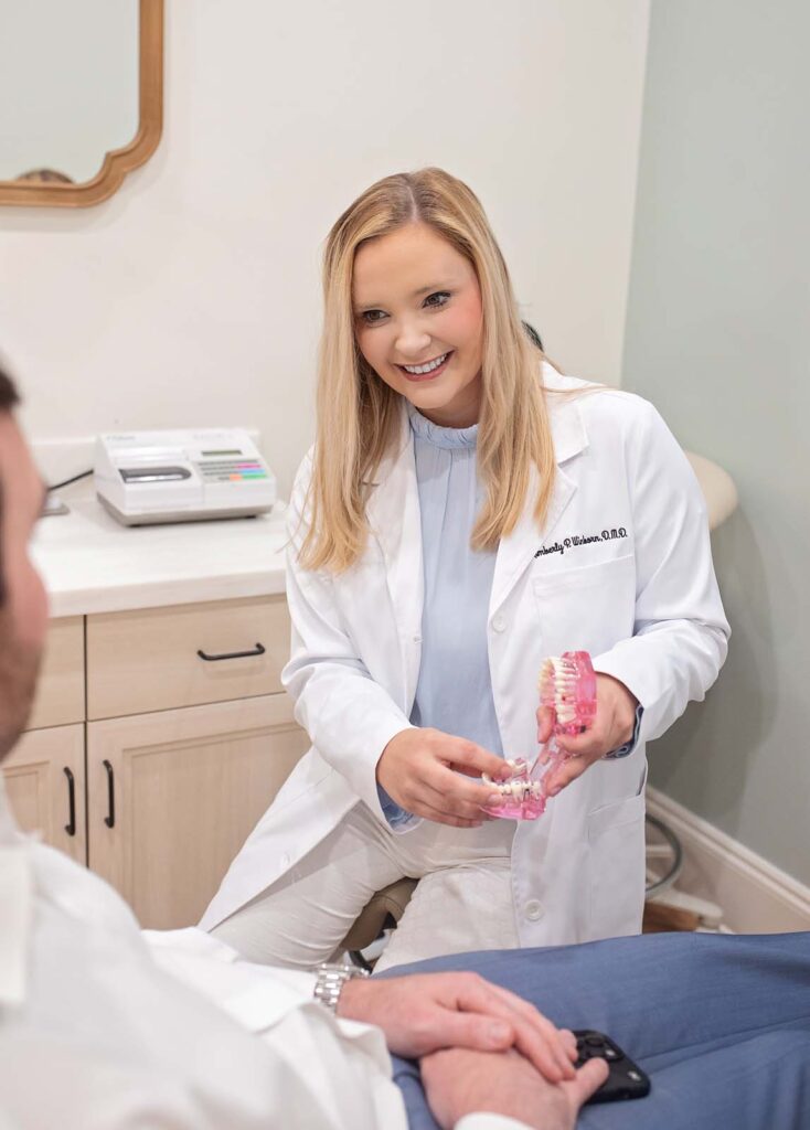 Dr. Winborn of Point Clear Dental, holding a dental model. They appear to be explaining or demonstrating something to a seated patient, who is partially visible in the foreground. The setting is a dental office, with a countertop, cabinets, and dental equipment visible in the background.