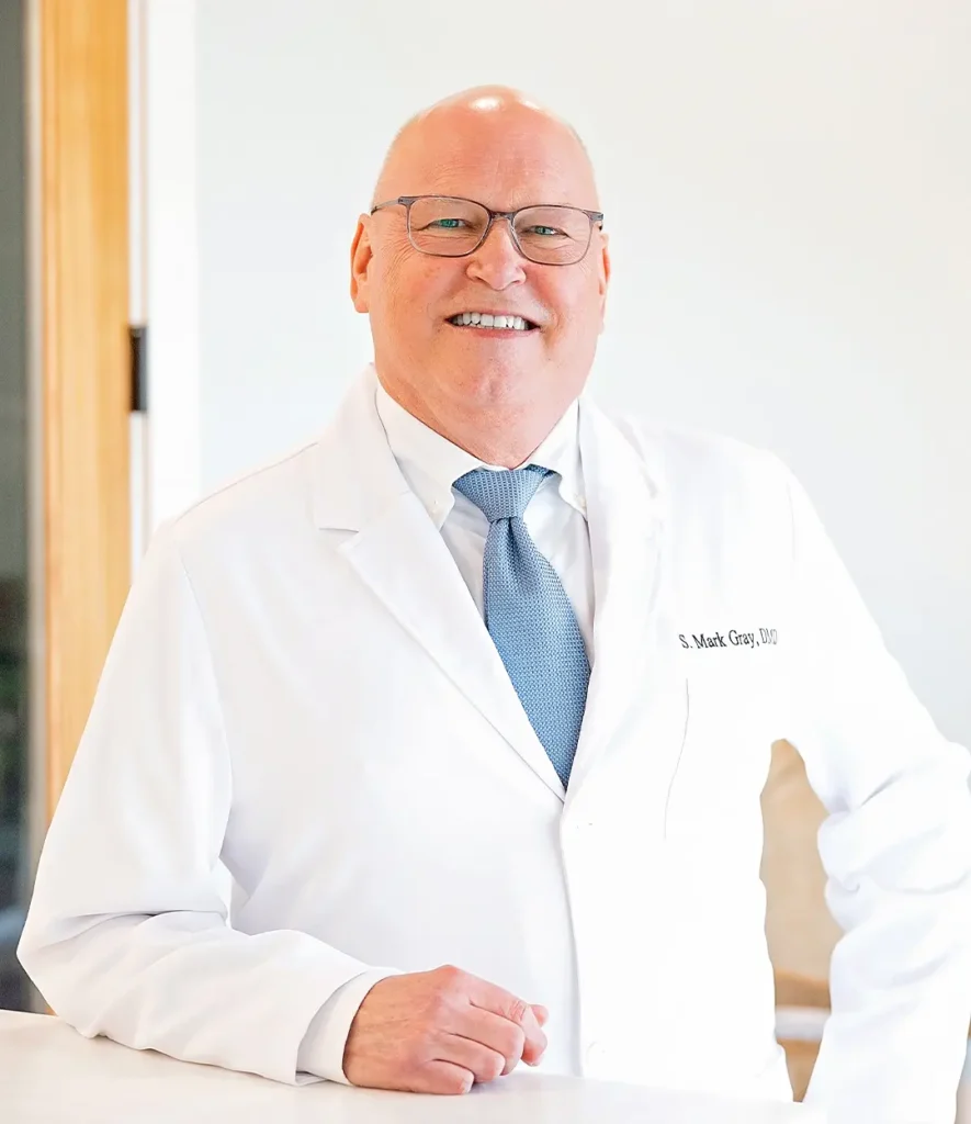 Smiling male dentist in white coat and glasses standing indoors, embroidered with 'S. Mark Gray, DDS
