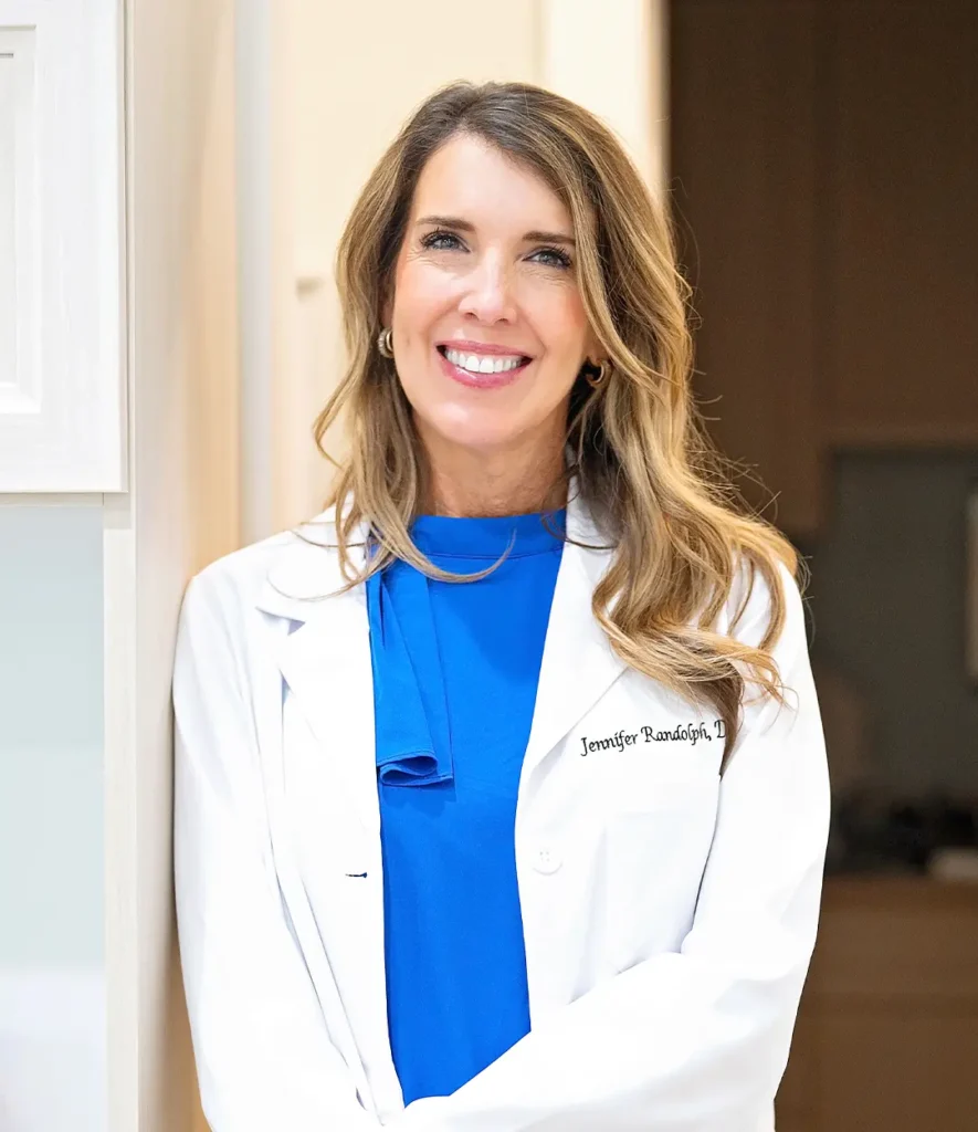 Smiling female dentist in white coat and blue blouse standing indoors, embroidered with 'Jennifer Randolph, D.
