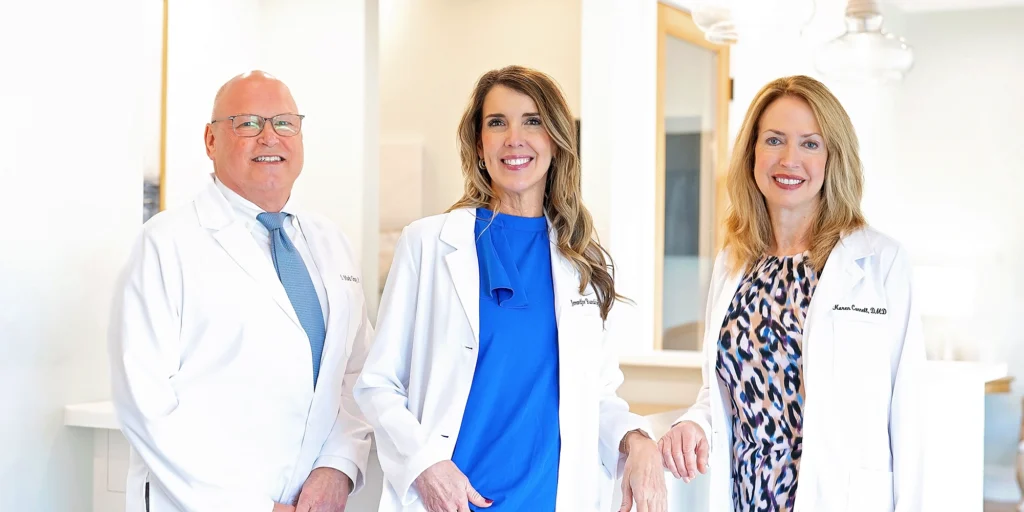 Three dental professionals in white lab coats standing together in a bright clinic setting.